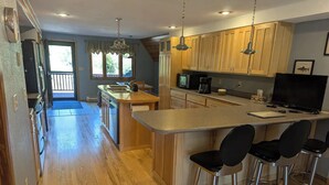 A modern kitchen with wooden cabinets, a central island with bar stools, a dining area with a table and chairs, and a large window providing natural light. - A-frame home on Trout Stream (Stoddard)