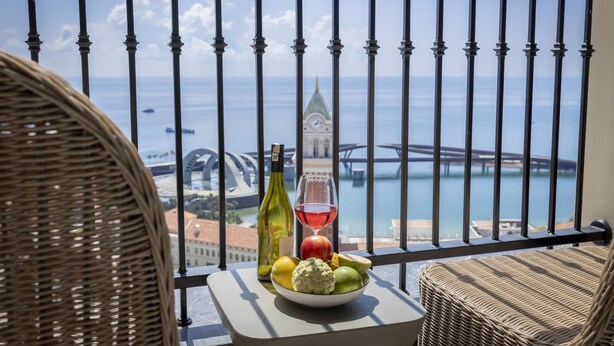 Een balkon met rieten stoelen, een tafel met fruit, een fles en een glas rode wijn, met uitzicht op een stadsgezicht met een brug en water.