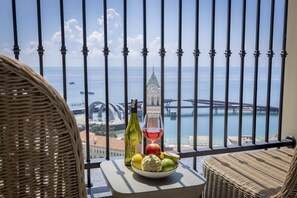 A balcony with wicker chairs, a table with fruits, a bottle, and a glass of red wine, overlooking a cityscape with a bridge and water. - Luxury Hillside Apartment Phu Quoc (Phu Quoc)