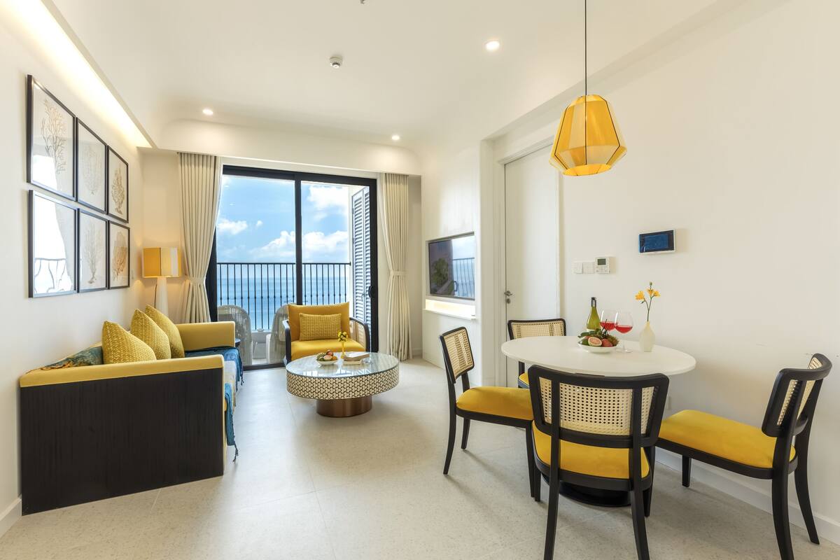 Une chambre d’hôtel moderne avec un balcon, une table à manger ronde entourée de chaises jaunes, un canapé avec des coussins jaunes et une grande fenêtre avec des rideaux.