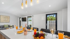 A modern kitchen with a central island, dining table, and chairs. There are various food items and drinks on the island, and a bottle of wine.