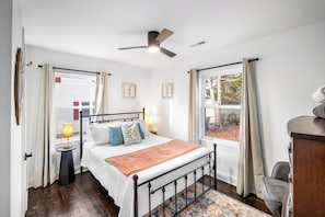 A bedroom with a bed, bedside lamp, ceiling fan, windows, and a view of the outdoors. - Buttercup Hideaway; Cozy oasis near WFU, Reynolda Gardens & Historic Graylyn (Winston-Salem)