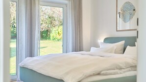 Une chambre à coucher avec un lit, une vue sur un jardin par une porte vitrée et une œuvre d’art encadrée au mur.