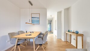 Un espace repas moderne avec une table en bois et des chaises à revêtement gris, un luminaire suspendu et une vue sur un couloir menant à une pièce dotée d’une grande fenêtre.