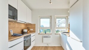 Une cuisine compacte avec des meubles blancs, un plan de travail en bois, un four et un micro-ondes encastré. Deux fenêtres laissent entrer la lumière naturelle et offrent une vue sur l’extérieur.
