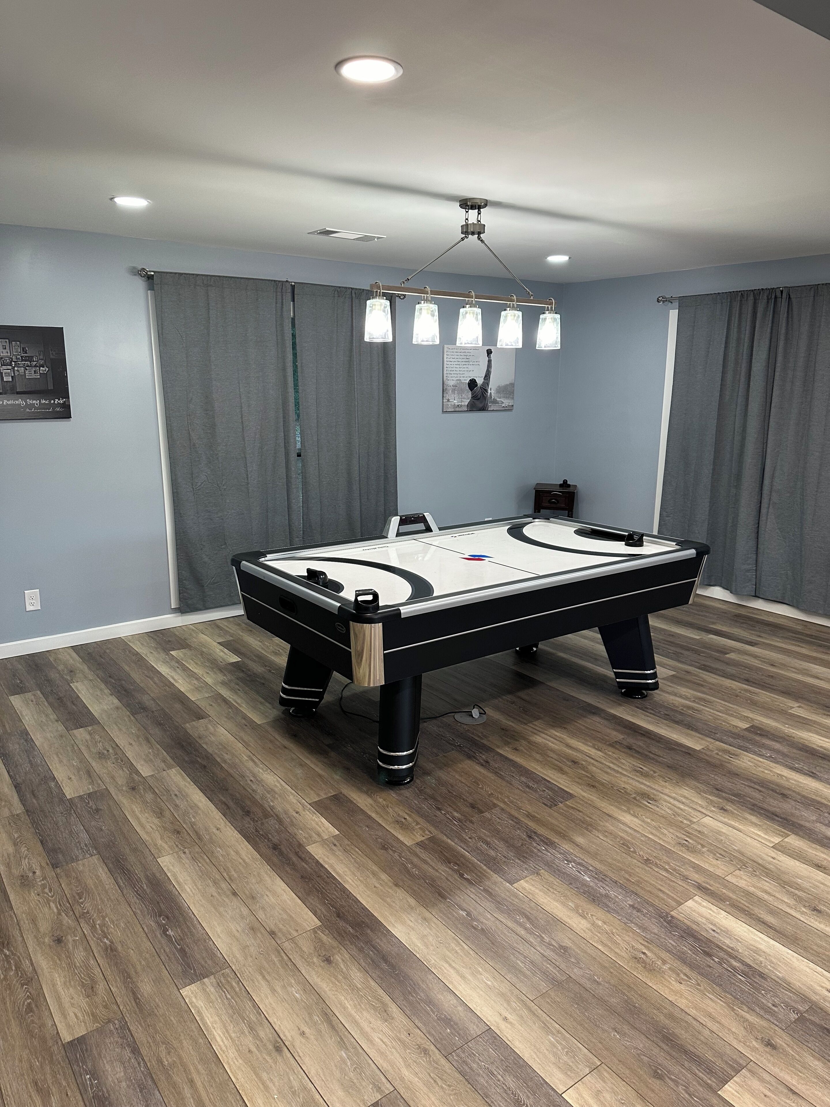 A pool table in a room with wooden flooring, grey curtains, and ceiling lights.