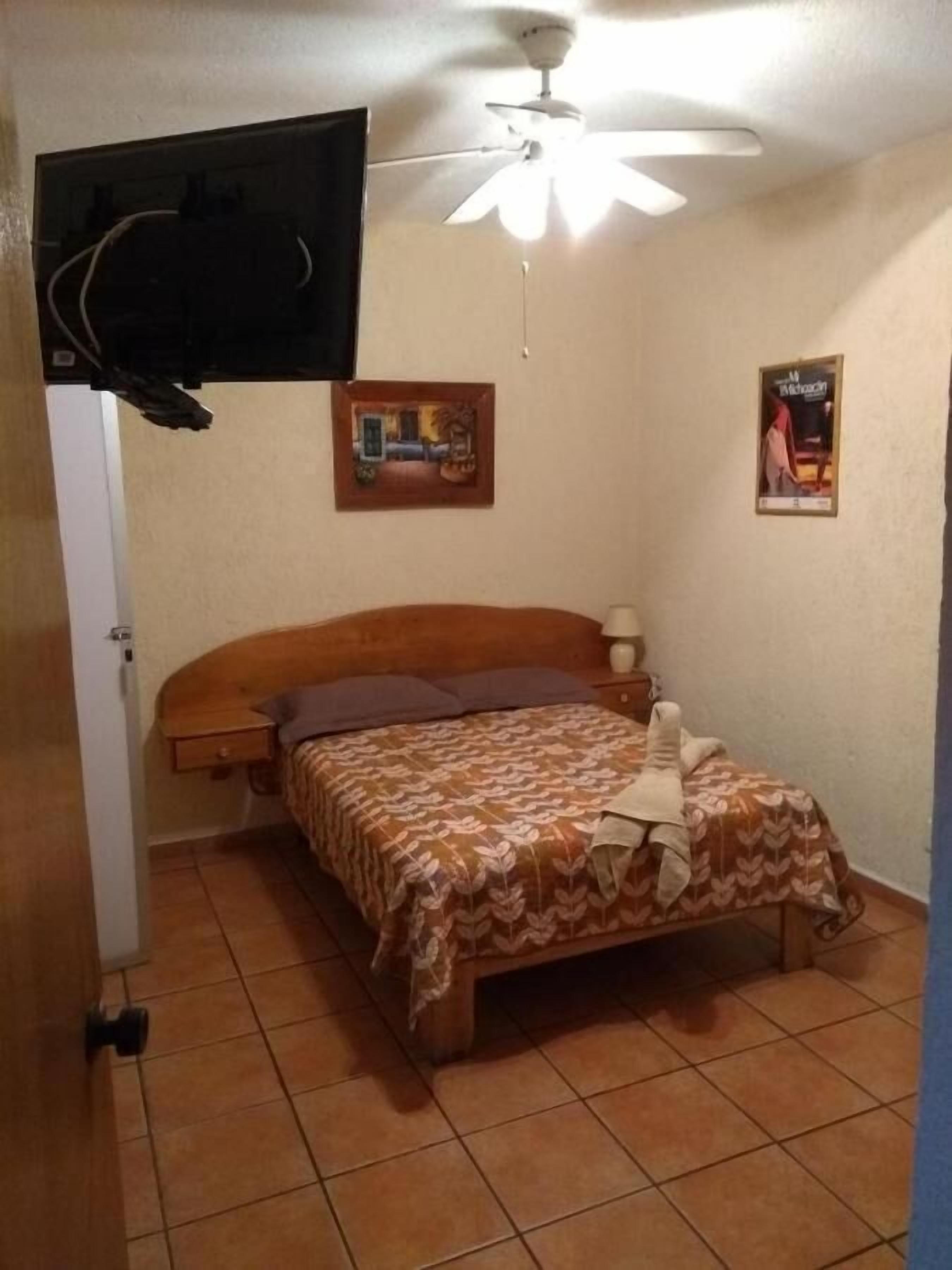 Un dormitorio con cama, ventilador de techo, televisión montada en la pared y dos cuadros enmarcados.
