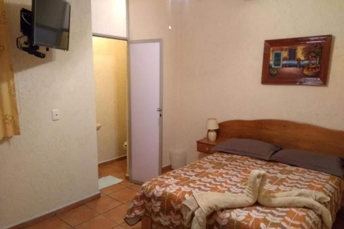 Un dormitorio con cama, mesita de noche, lámpara, televisor montado en la pared y puerta que da a otra habitación.