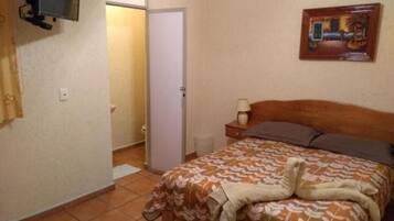 Quarto com cama, mesa de cabeceira, abajur, televisão fixada na parede e porta que dá para outro cômodo.