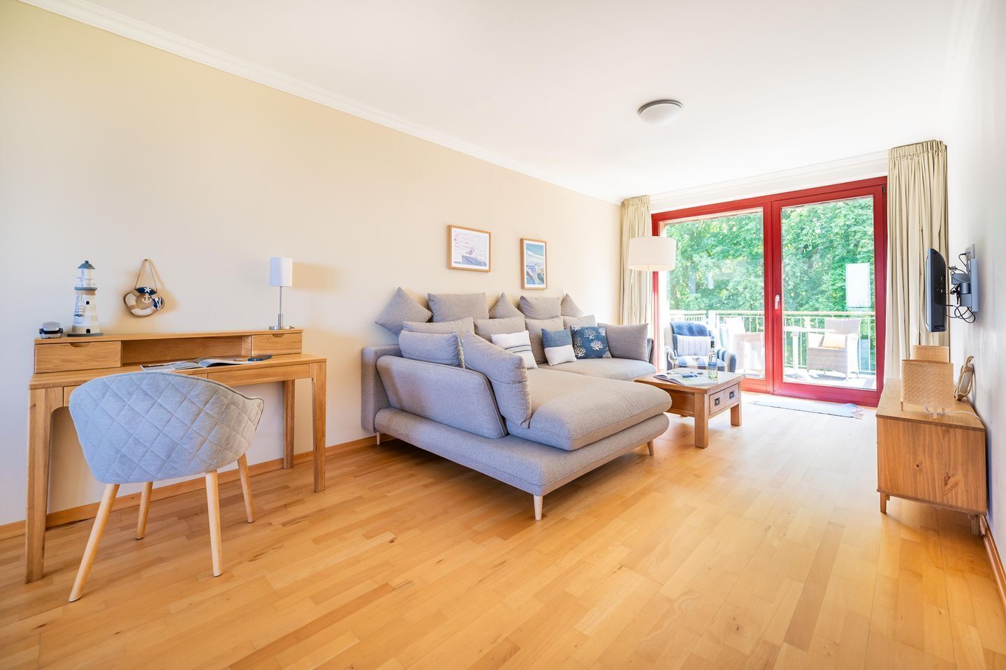 Un salon moderne avec un canapé, un bureau et une chaise. Il y a un balcon avec une porte rouge et un téléviseur fixé au mur.