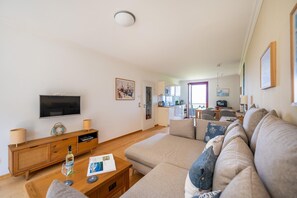 A modern living room with a large sofa, a wooden coffee table, and a flat-screen TV. A kitchen is visible in the background with a dining area.