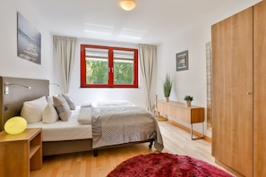 A bedroom with a bed, bedside tables, a wardrobe, a window with curtains, and a view of greenery outside.