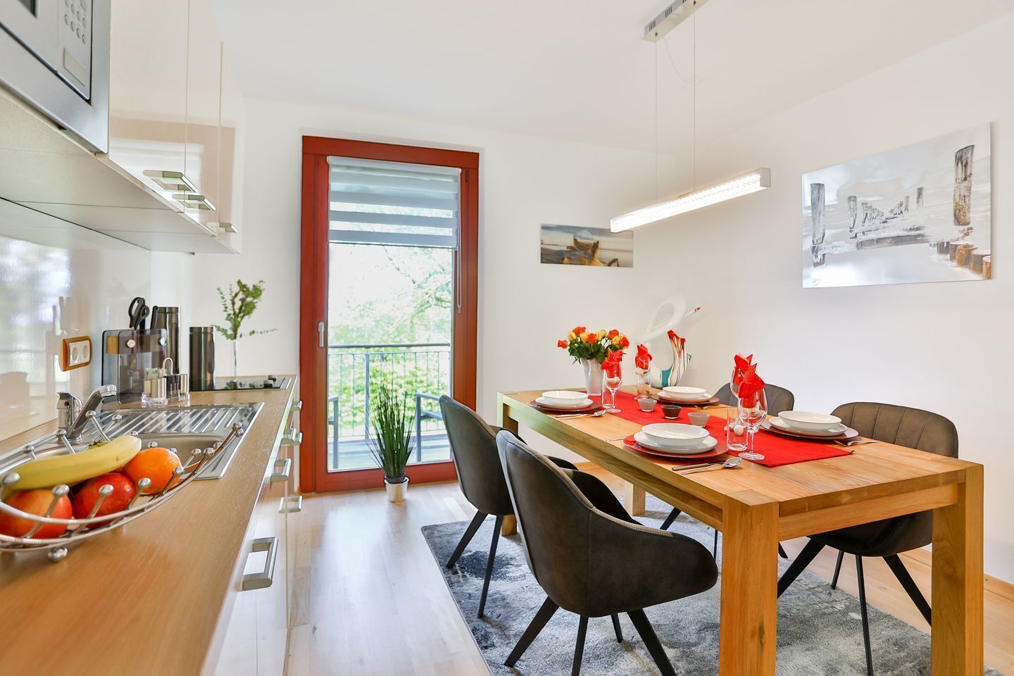 A modern kitchen with a dining area, featuring a wooden table, black chairs, and a view of the outdoors through a large window.