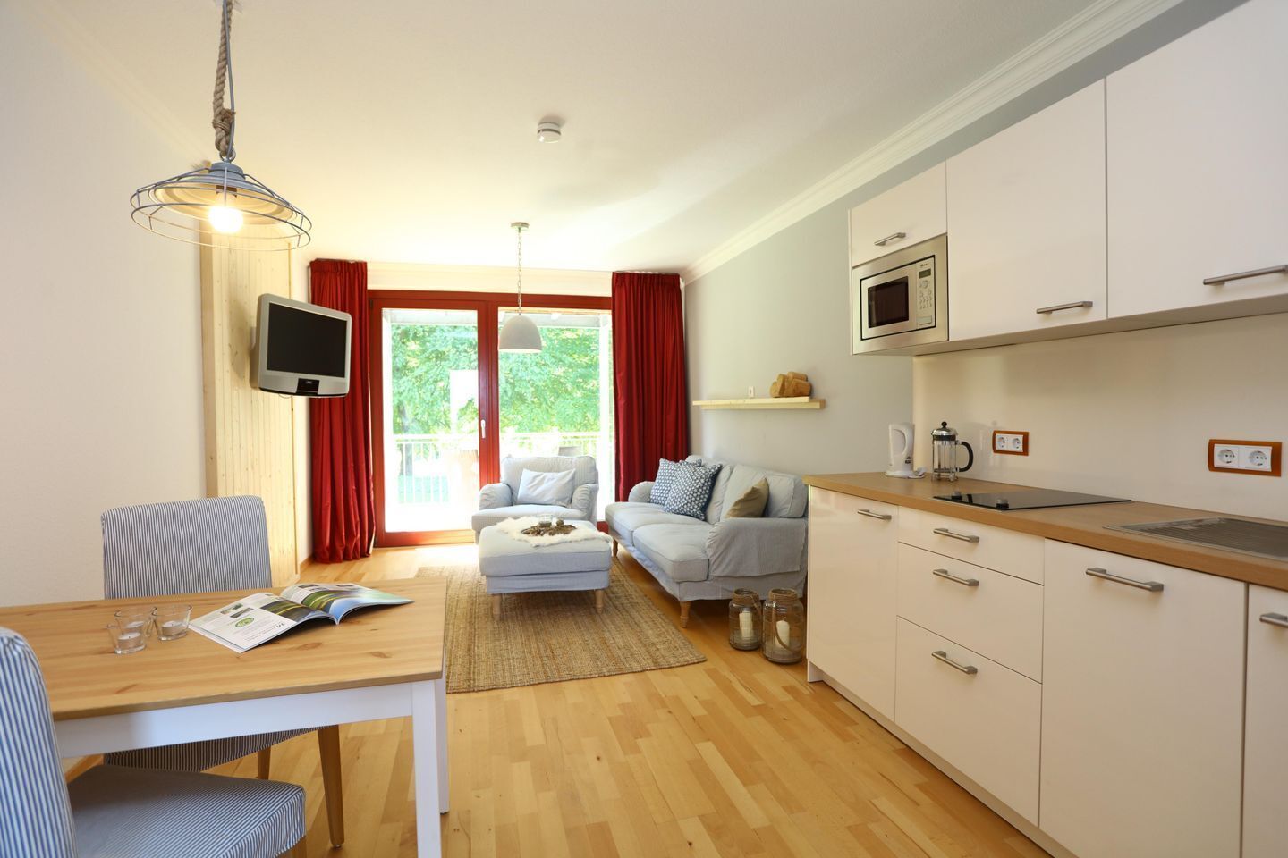 A modern living room with a sofa, a dining table with chairs, a kitchen area with cabinets and a microwave, and a television.