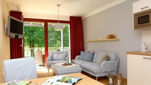 Una sala de estar acogedora con sofá, reposapiés y una mesa de comedor con una revista y vasos. Un televisor está montado en la pared y hay una zona de cocina con un microondas y un estante encima.