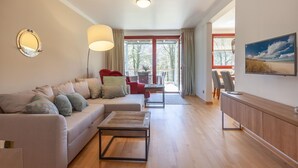 A spacious living area with a sofa, a coffee table, a red armchair, and a dining table. There is a large window with curtains, a painting on the wall, and a ceiling light.
