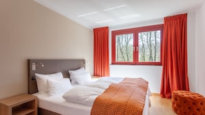 Un dormitorio moderno con una cama grande, una mesita de noche, una ventana con cortinas rojas y vistas a árboles.