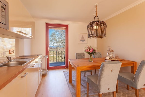 A modern kitchen with a built-in oven, a dining area with a wooden table and grey chairs, and a balcony with a red door.