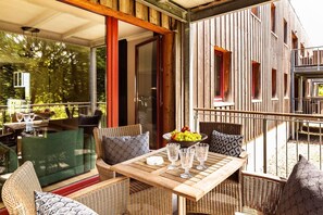 Un espace extérieur aménagé en terrasse, avec des meubles en osier, une table sur laquelle se trouve un saladier rempli de fruits et une rambarde en verre.