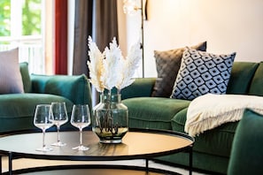 Un divano verde con cuscini decorati, un vaso di vetro con fiori bianchi e tre bicchieri da vino su un tavolo rotondo.