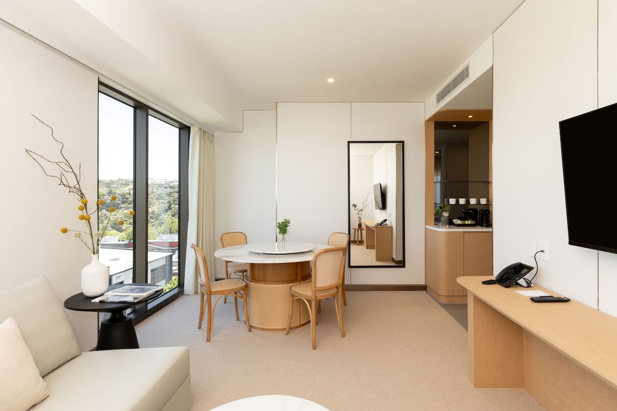 Une chambre d’hôtel moderne dotée d’une grande fenêtre, d’une table à manger ronde avec des chaises, d’un canapé, d’un bureau avec un téléphone et d’un téléviseur à écran plat.
