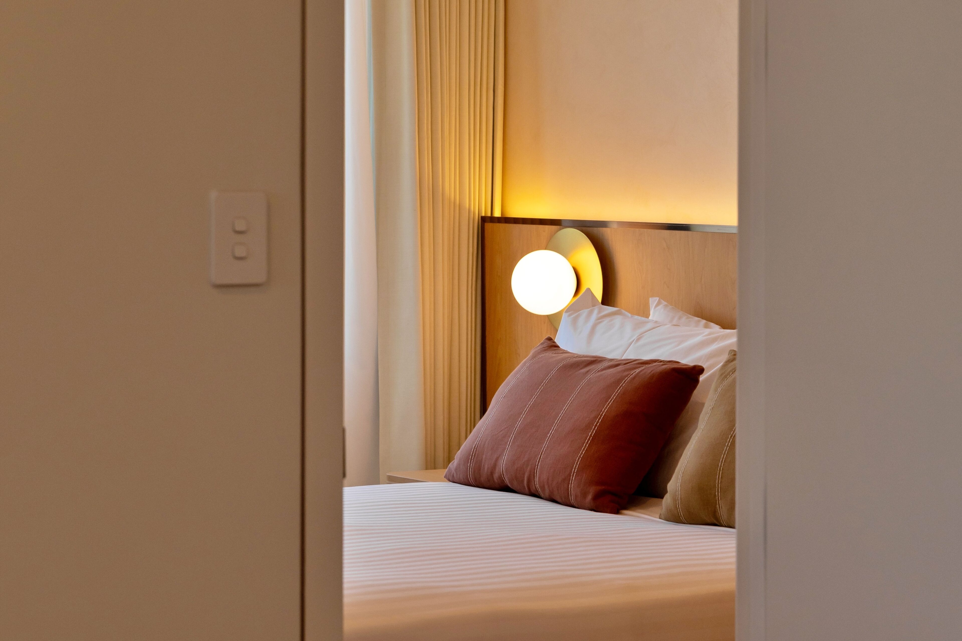 A hotel room with a neatly made bed, a headboard with a round light, and a visible power outlet on the wall.