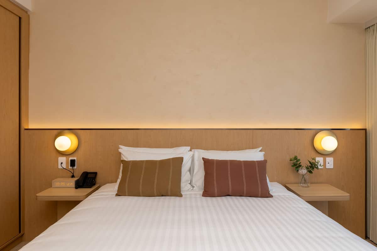 Une chambre d’hôtel avec un grand lit, deux tables de chevet, une petite table, une horloge murale et deux lampes fixées au mur.