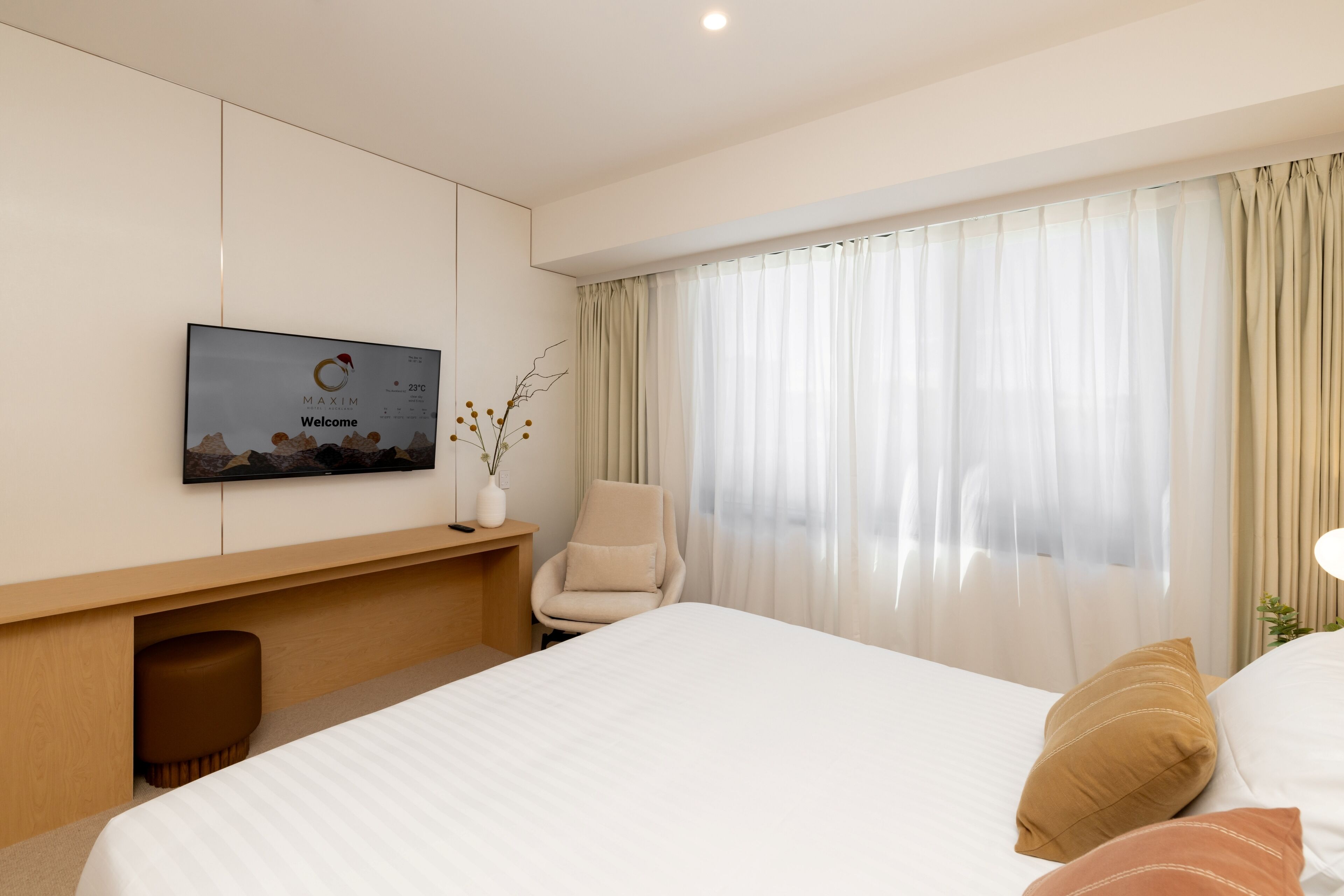 A modern hotel room with a large bed, a flat-screen TV mounted on the wall, a comfortable armchair, and a large window with sheer curtains.
