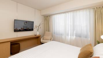 A modern hotel room with a large bed, a flat-screen TV mounted on the wall, a comfortable armchair, and a large window with sheer curtains.