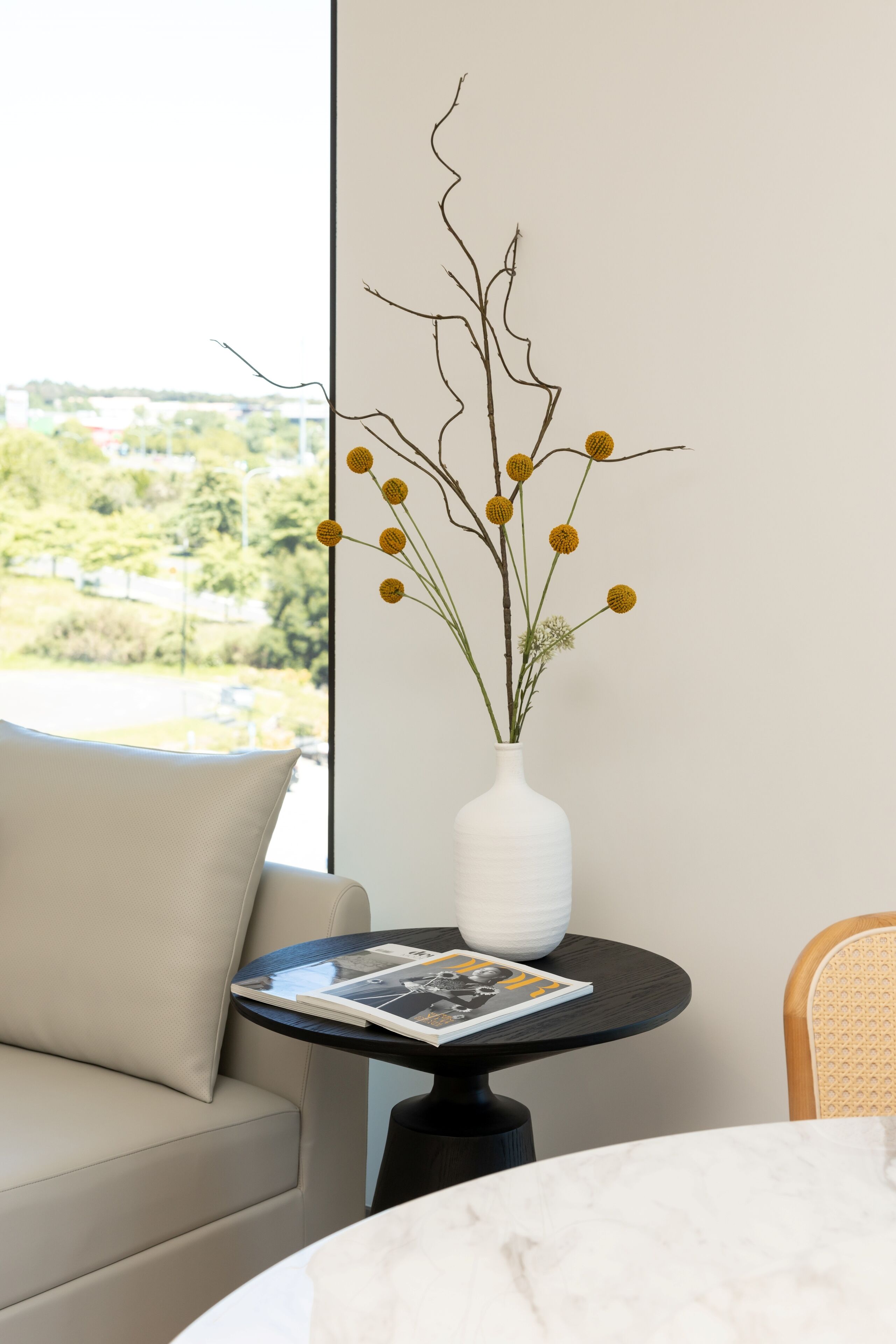 Una sala de estar moderna con un sofá color beige, una mesa redonda con una revista y un jarrón blanco con flores amarillas.