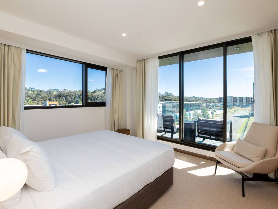 A modern hotel room with a large bed, a comfortable armchair, and a balcony with a city view.