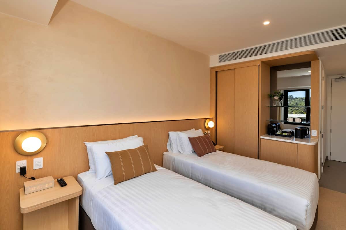 Une chambre d’hôtel avec deux lits, une table de chevet avec un téléphone et une horloge numérique, une petite kitchenette équipée d’une machine à café, et un grand miroir au-dessus du comptoir.