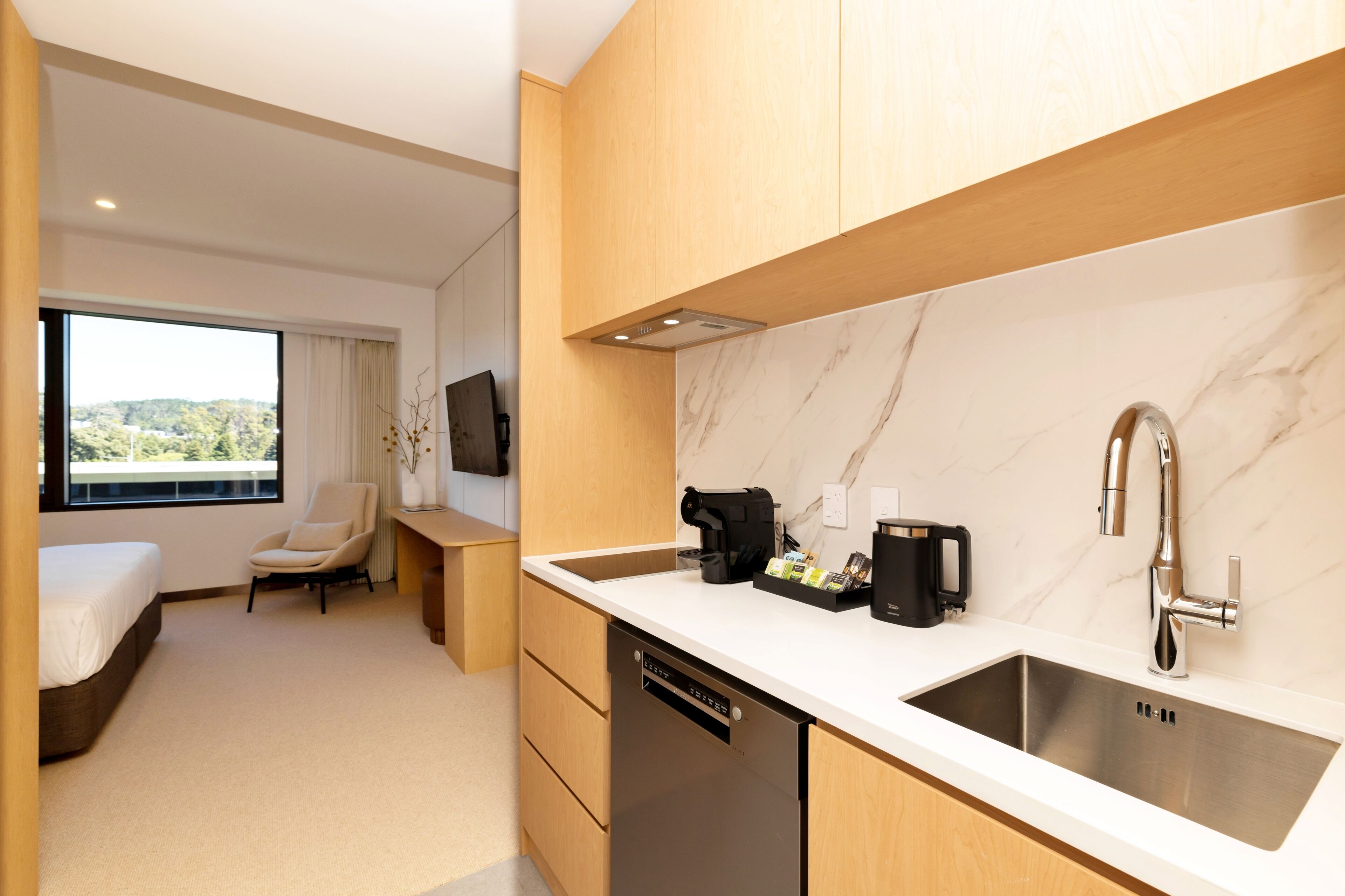 A modern hotel room with a kitchen area, a bed, a chair, and a large window offering a view of the outdoors.