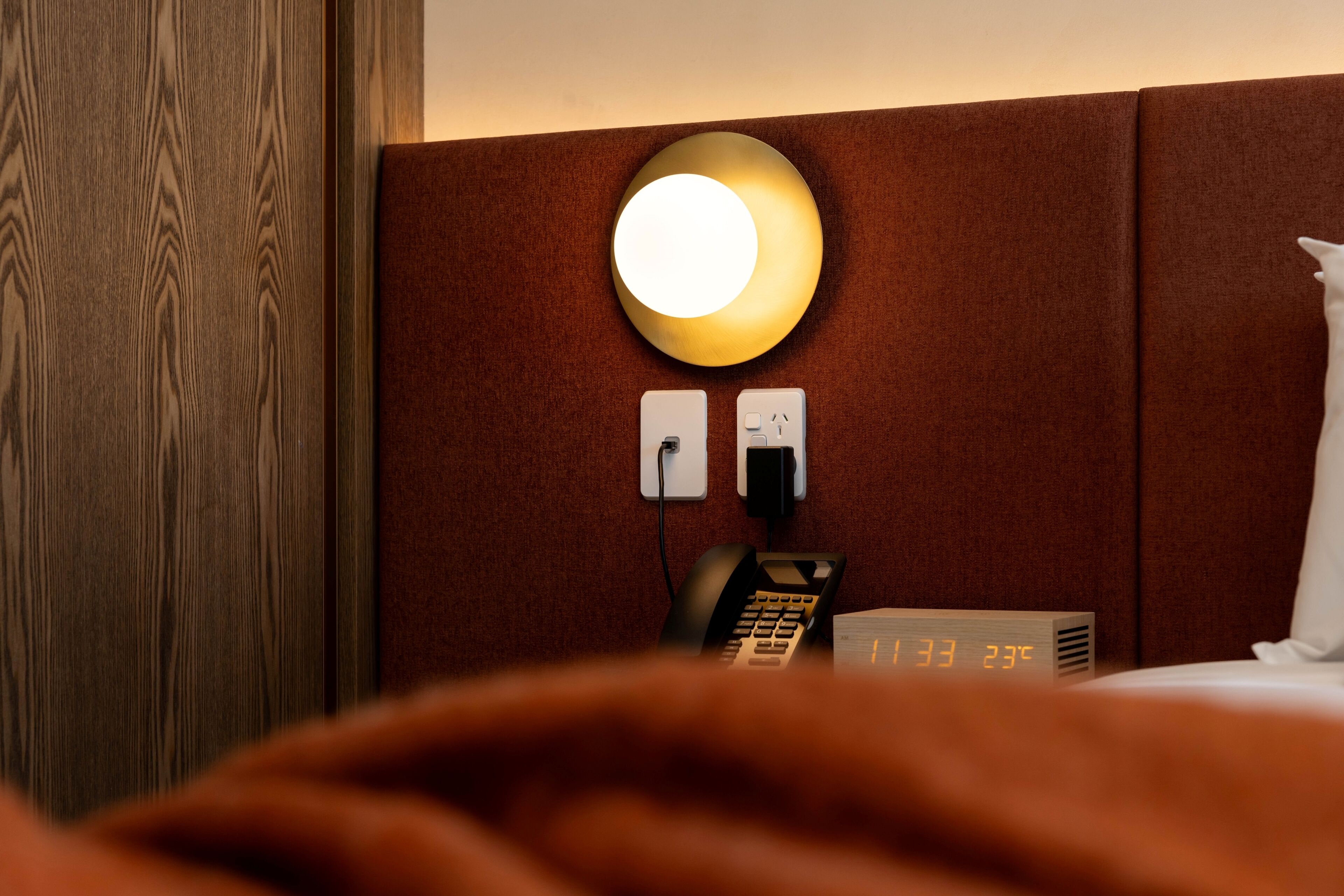 A hotel room with a wooden headboard, a bedside lamp, a telephone, and a digital clock displaying the time and temperature.