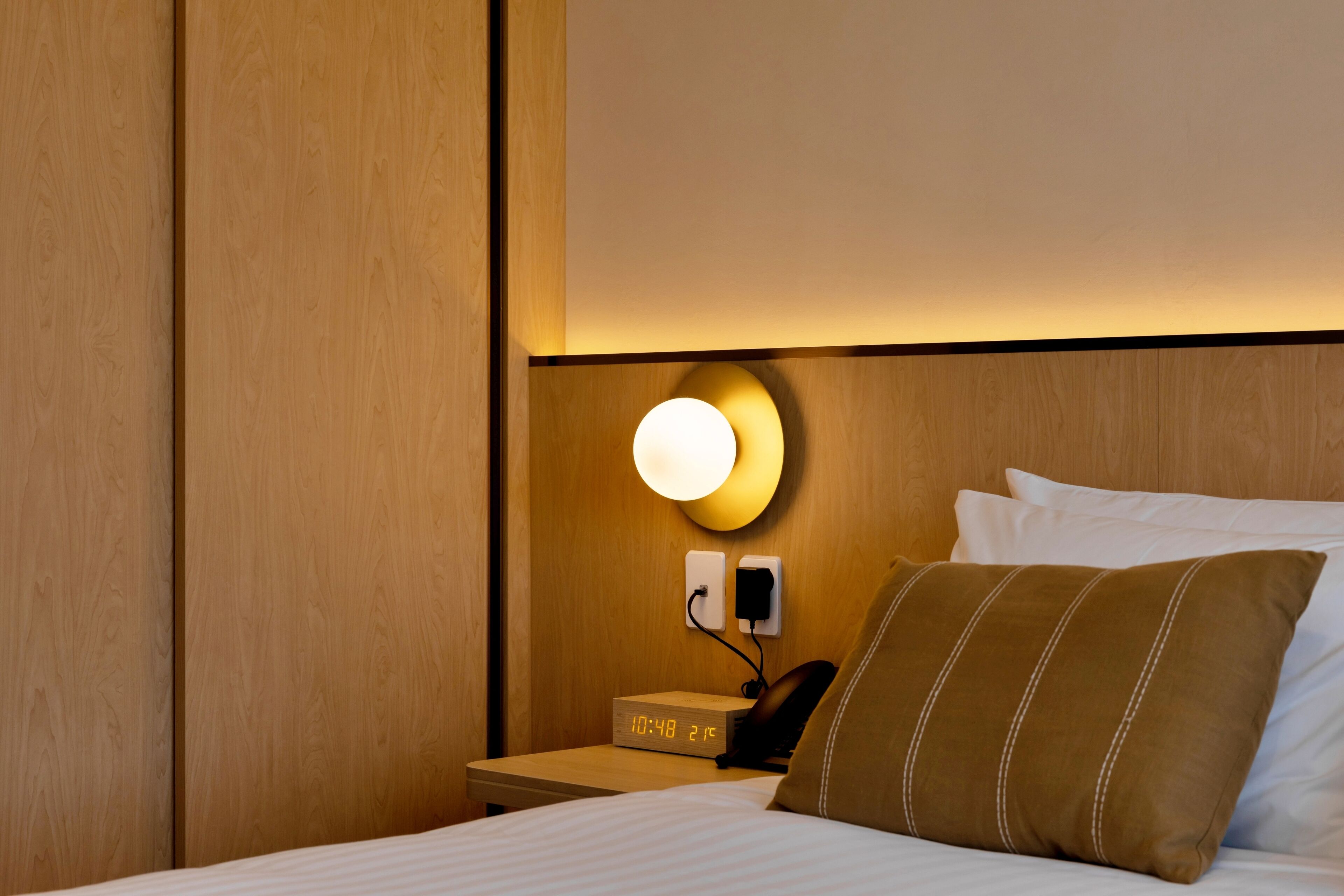 Habitación de hotel con cabecera de madera, mesita de noche con reloj y teléfono, luz empotrada en la pared y cama con ropa blanca y almohada marrón.