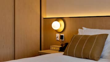 Habitación de hotel con cabecera de madera, mesita de noche con reloj y teléfono, luz empotrada en la pared y cama con ropa blanca y almohada marrón.