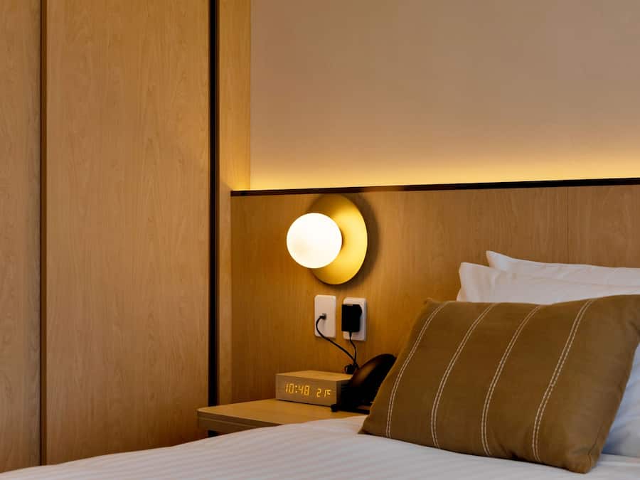 Habitación de hotel con cabecera de madera, mesita de noche con reloj y teléfono, luz empotrada en la pared y cama con ropa blanca y almohada marrón.