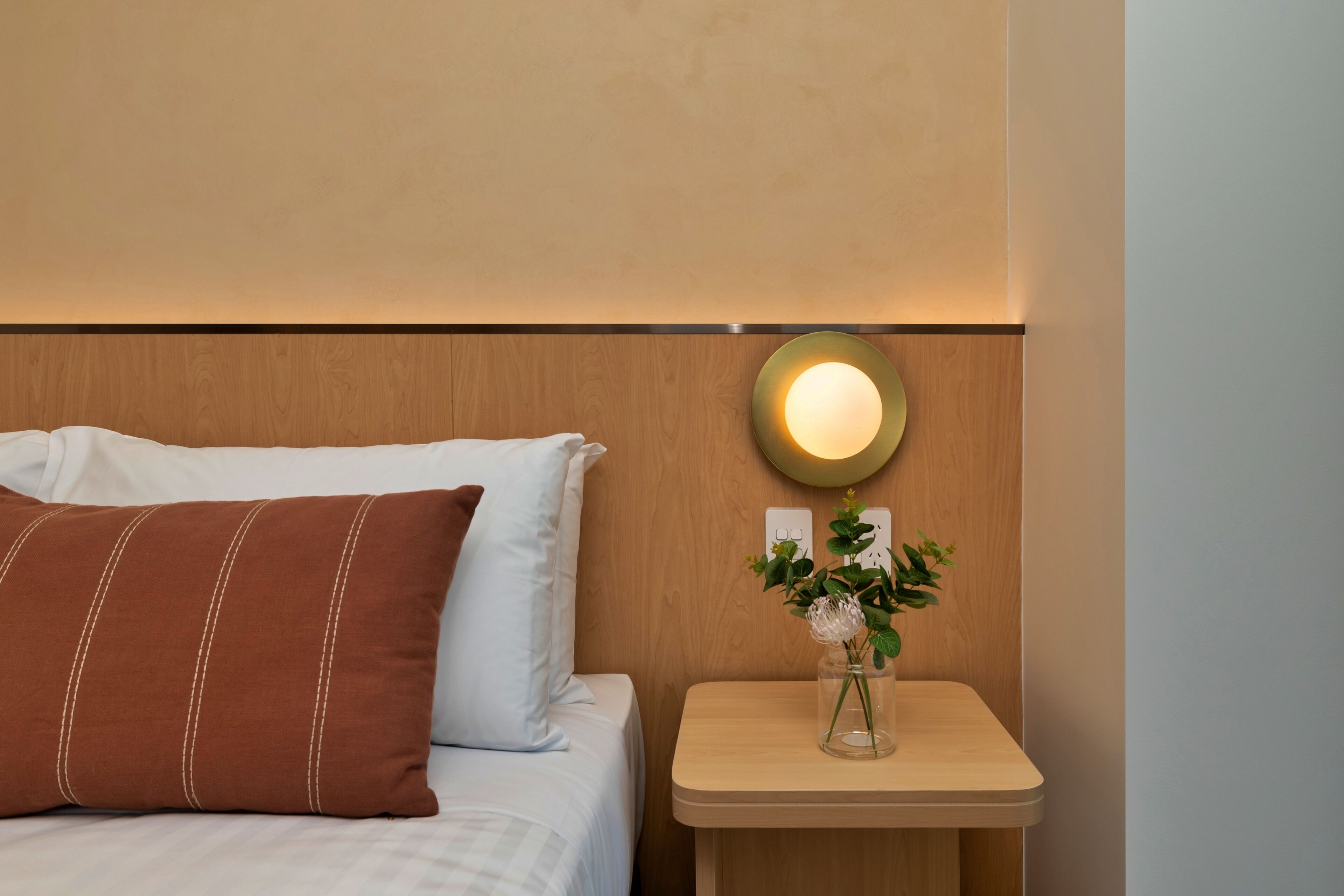 A neatly made bed with a brown pillow and a white pillow, a bedside table with a vase of flowers, and a wall-mounted light fixture.