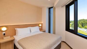 Una habitación de hotel moderna con una cama grande, una mesita de noche con una lámpara y un ventanal con vistas al exterior.