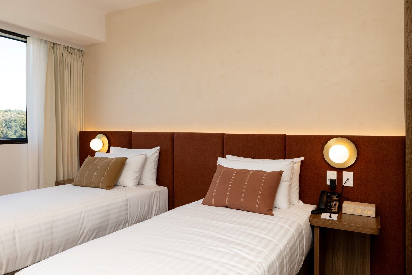 Habitación de hotel con dos camas, cada una con sábanas blancas y almohadas marrones. Hay lámparas de noche con luces redondas, una mesita de noche de madera con un teléfono y un reloj digital, y una ventana con cortinas.