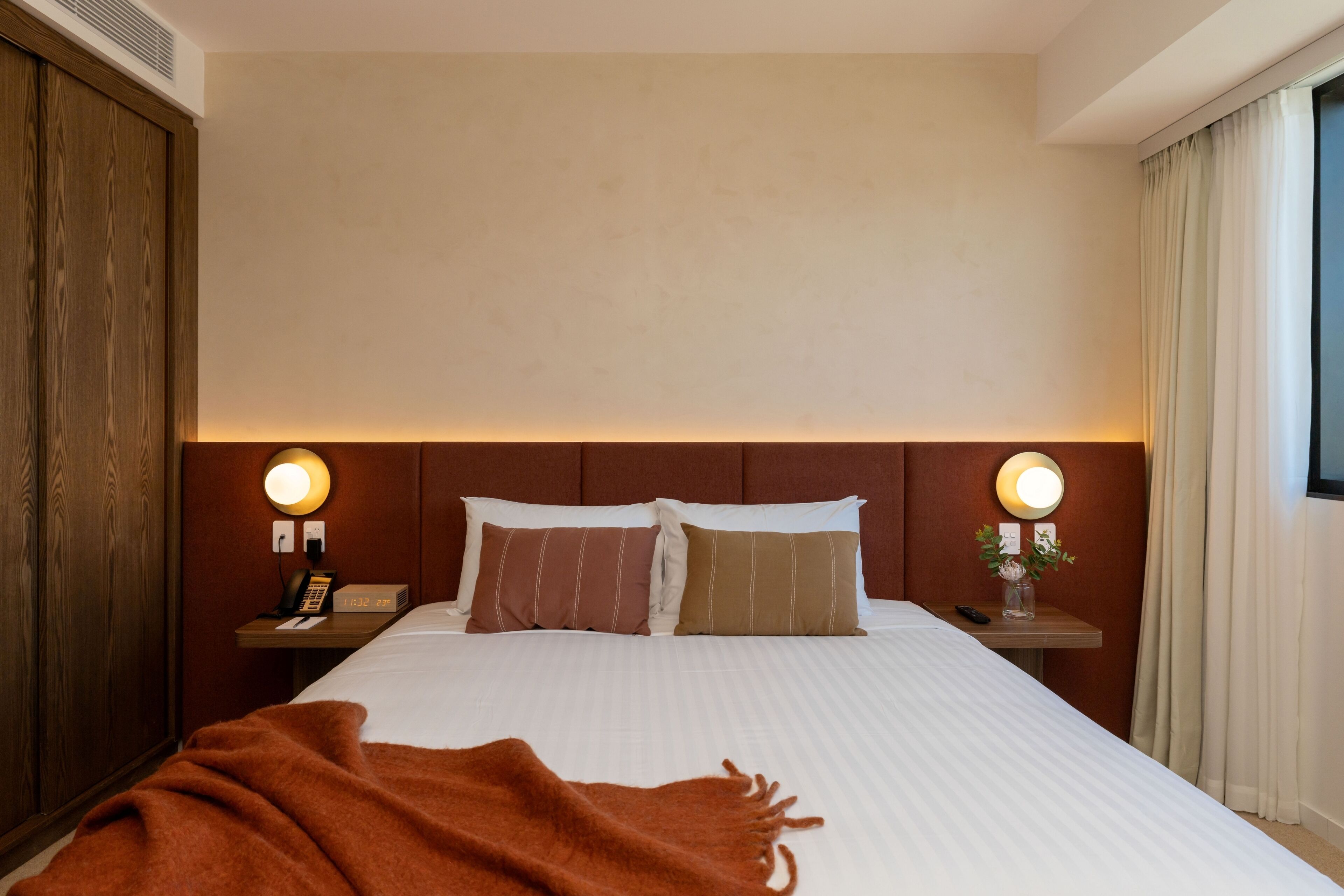 A hotel room with a large bed, two bedside tables, a wooden wardrobe, and a window with curtains.