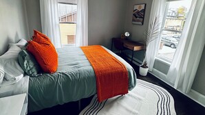A bedroom with a bed, a desk, a chair, a potted plant, and a window with curtains.