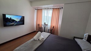 Een hotelkamer met een bed, een televisie aan de muur, een kleintje tafeltje met stoelen en een raam met gordijnen.