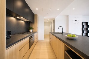 A modern kitchen with a black countertop, wooden cabinets, a built-in stove, and a sink. There is a clock on the wall and a plant on the counter. - The Evelyn Port Melbourne by Ready Set Stay - WIFI, linen, towels, brand new (Port Melbourne)
