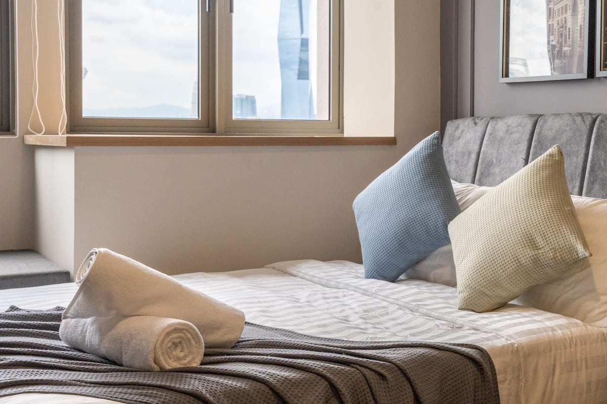 A neatly made bed with a rolled towel, a gray upholstered headboard, and a view of buildings through a window.