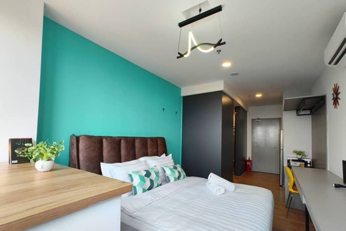 A modern hotel room with a teal accent wall, a wooden desk, a bed with white bedding, and a unique ceiling light fixture.