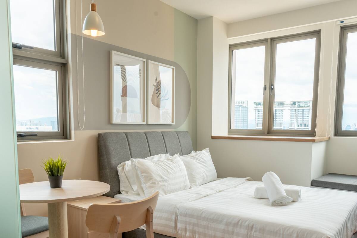 A hotel room with a bed, a desk, a chair, a potted plant, and large windows offering a city view.