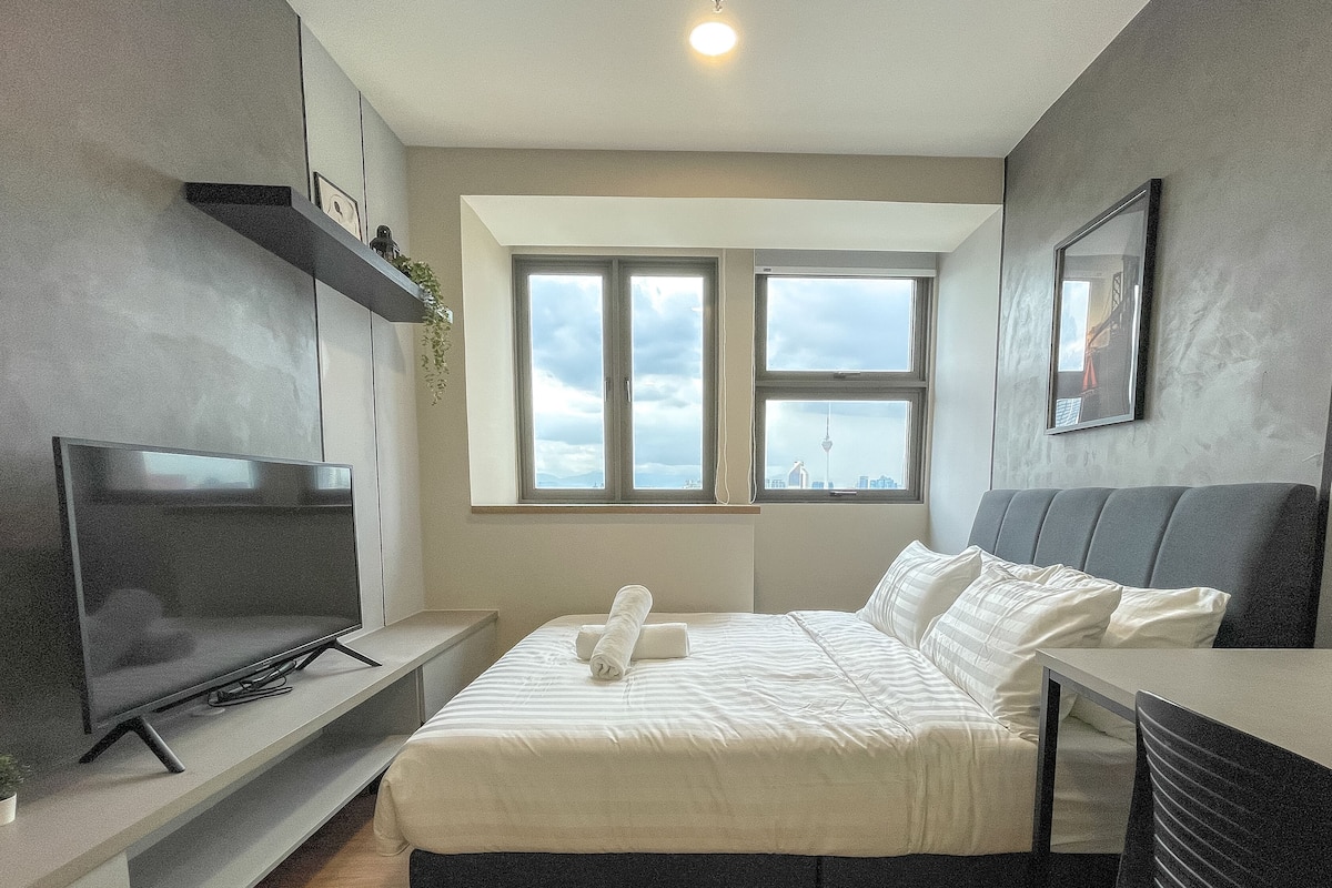 A modern bedroom with a bed, a TV, a desk, and a window with a city view.
