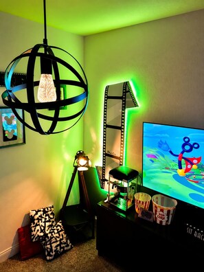 Una habitación con un escritorio, un monitor de computadora mostrando un dibujo animado, una lámpara de película y una lámpara globo.