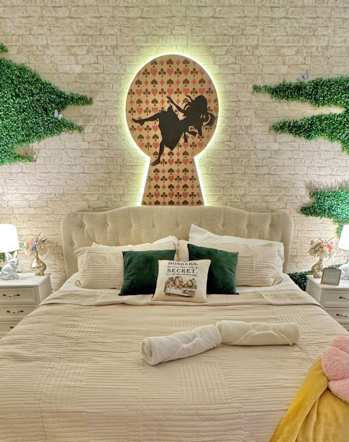 A bedroom with a tufted headboard, a bed with a quilted bedspread, pillows, a folded towel, and a wall-mounted decorative piece.